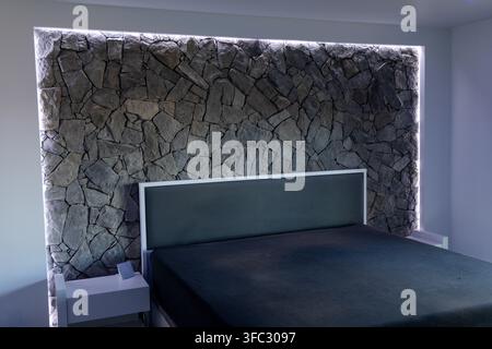 A contemporary bedroom features a dramatic stacked stone wall behind the bed, enhanced by subtle LED backlighting for ambiance Stock Photo