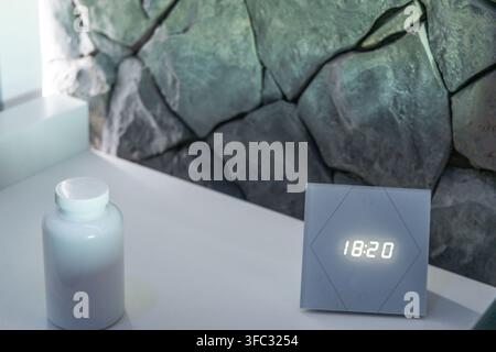 A minimalist digital clock showing 18:20 sits beside a white opaque bottle on a clean white surface with a stone wall background Stock Photo