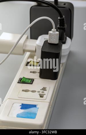 Overloaded white power strip with multiple electrical plugs, adapters, and a USB charging cable plugged into the sockets. Stock Photo