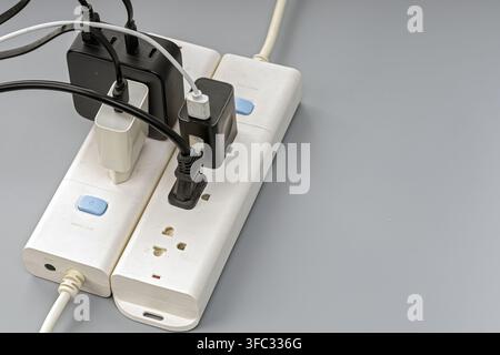 White power strip crowded with black chargers, adapters, and tangled cables, showing risk of electrical overload and fire hazard. Stock Photo