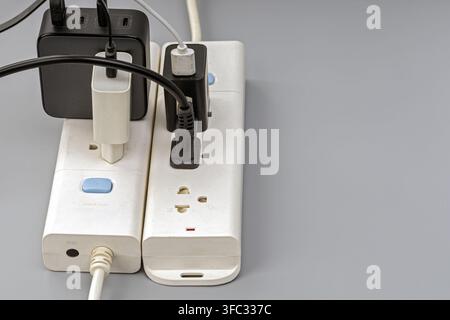 Overloaded power strip with tangled cables and multiple chargers plugged in, showing a risky electrical setup on a gray surface. Stock Photo