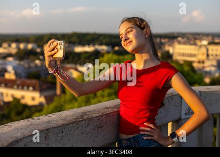 Young Woman Taking Smartphone Selfie at Golden Hour. Young woman in a red top taking a smartphone selfie on a rooftop at golden hour with a blurred Stock Photo