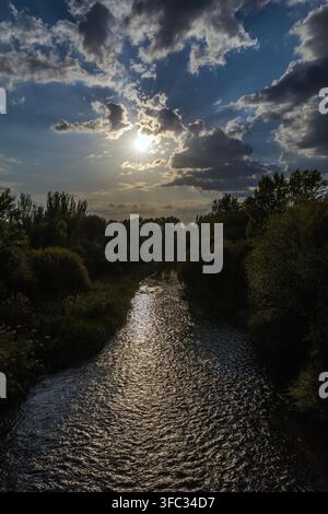 A scenic river flows through lush green banks under a dramatic sunset sky with sun rays shining through clouds in Spain. Stock Photo