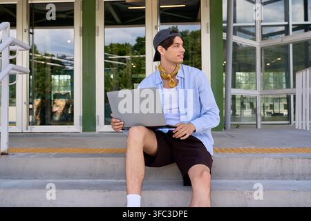 A young man in a blue shirt sits outdoors, working on his laptop on campus Stock Photo