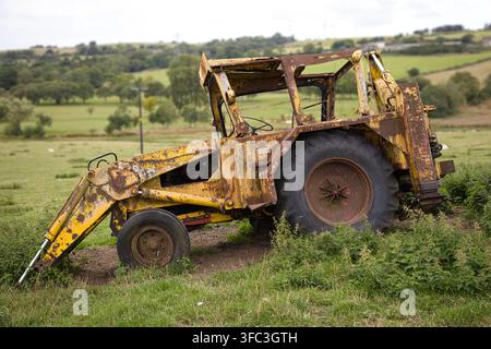 Profile view of an obsolete rusted yellow backhoe tractor left to decay in a idyllic rolling countryside pasture on an overcast day. Stock Photo