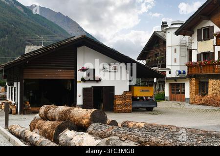 Wooden logs stacked in front of a local sawmill workshop with traditional alpine chalets and mountains behind, Cogne, Italy Stock Photo