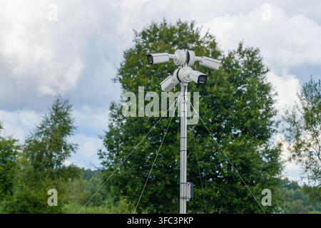 Four CCTV security cameras mounted on stabilized outdoor pole provide multi-directional video surveillance against green landscape and cloudy sky Stock Photo
