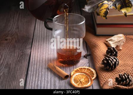 Pouring hot black tea into a glass cup. Rustic autumn composition with dried orange slices, cinnamon, and pine cones on a wooden table with copy space Stock Photo