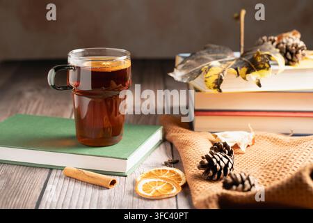 Glass cup of hot spiced tea placed on a green book. Cozy autumn aesthetic with stacked books, dry leaves, pine cones, and cinnamon on wooden desk Stock Photo