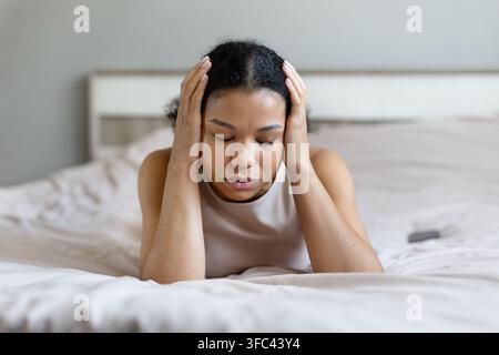 Young Black woman lies on bed with eyes closed, holding head with both hands, suffering from headache or exhaustion at home, appearing stressed in her Stock Photo