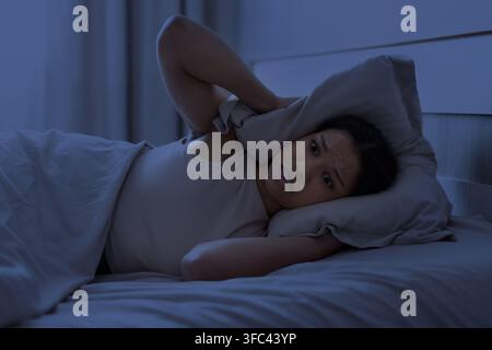 Young Black woman lying awake at night with pillow over her ear Stock Photo