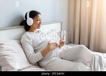 Smiling pregnant Black woman with smartphone relaxing on bed wearing wireless headphones Stock Photo