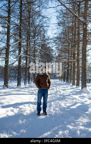 Young man standing alone on a snowy path among tall trees, looking sad and thoughtful, conveying loneliness, solitude, dreams and winter melancholy. V Stock Photo