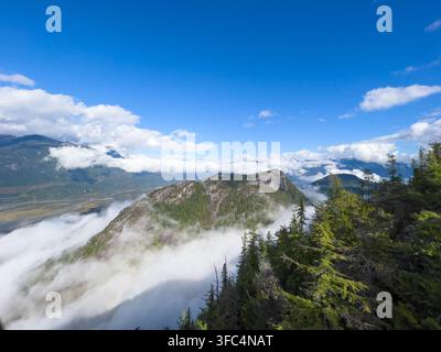 A rugged granite mountain peak rises above a thick layer of cloud, framed by forested ridge lines under a clear blue sky. Stock Photo