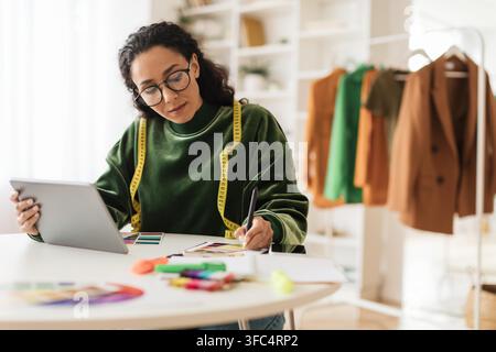 Young Hispanic Female Designer Working With Tablet And Sketches At Fashion Studio, Creative Concept Stock Photo
