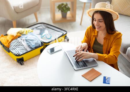 Happy Young Latina Woman Using Laptop While Preparing For Trip, Home Office Setting Stock Photo