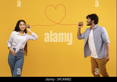 Happy South Asian Couple Talking Via Tin Can Phone, Yellow Background, Love Concept Stock Photo