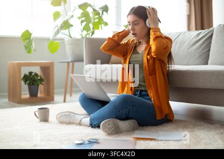 Surprised Young Hispanic Woman Wearing Headphones At Home Using Laptop, Online Concept Stock Photo