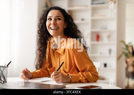 Happy Young Woman With Curly Hair Smiling While Taking Notes In Modern Office, Business Concept Stock Photo