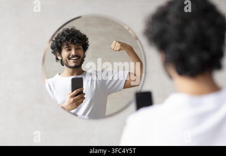Smiling South Asian Male Flexing Arm Muscles In Front Of Mirror, Home Workout Concept Stock Photo