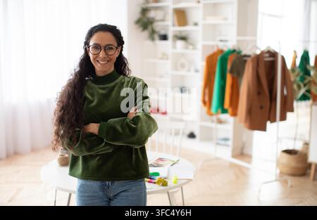 Smiling Hispanic Woman Standing Confidently In Workspace With Colorful Clothing Rack, Business Concept Stock Photo