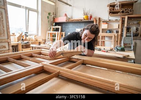 Attentive female carpenter carefully examines parts of old wooden window frame before restoration. Stock Photo