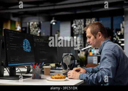 Engineer utilizing AI on computer to optimize performance while eating croissant. Worker in tech company office taking snack break while training machine learning models to perform automatic updates Stock Photo