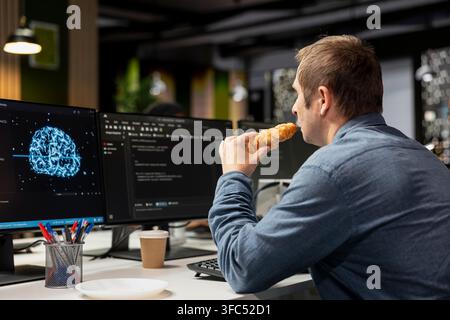 Programmer in tech startup office eating croissant and automating repetitive tasks using AI script on PC. Worker taking snack break, waiting for AI assistant to speed up feature implementation Stock Photo