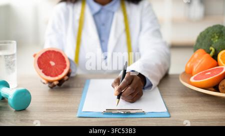 Black Female Nutritionist Holding Grapefruit And Taking Notes At Office, Weight Loss Concept Stock Photo