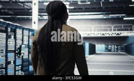 Woman walking through data center server rows, checking equipment for system stability. IT professional overseeing server farm, monitoring infrastructure mainframes, camera A Stock Photo