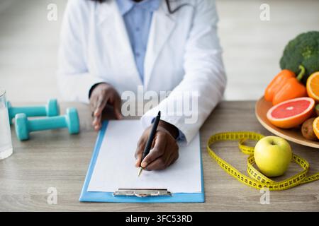 Black Female Nutritionist Writing Meal Plan With Healthy Foods On Table, Clinic Healthcare Concept Stock Photo