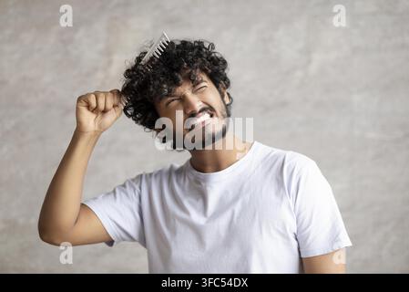 Young Man With Curly Hair Combing Hair With Comb At Home, Frustrated Expression Stock Photo