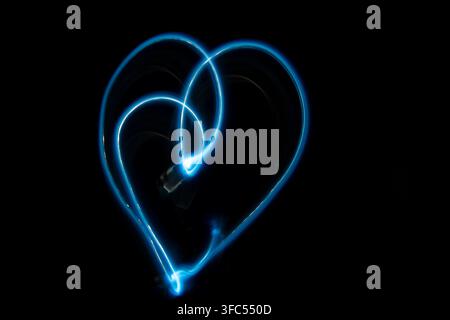 A romantic and modern long exposure photograph capturing a glowing neon blue light trail that forms the shape of a heart in darkness. Stock Photo