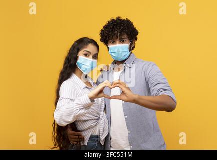 Couple Hugging And Making Heart Shape With Hands, Wearing Masks, Yellow Background Stock Photo