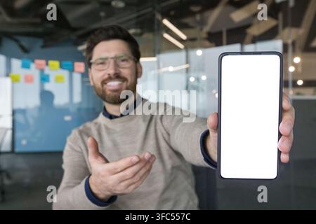 Smiling man in a modern office presenting a mobile phone with a blank white screen, extending his arm and gesturing to the device ideal for app, tech or business mockups Stock Photo