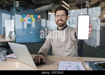 Young businessman in modern office smiling as he holds a smartphone with blank screen toward camera for app mockup, working at laptop surrounded by documents and charts Stock Photo