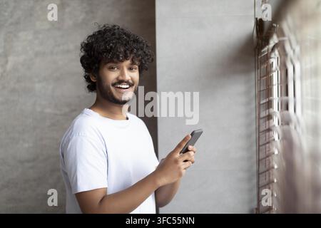 Smiling Young Asian Man Using Smartphone, Standing By Window At Home, Lifestyle Concept Stock Photo
