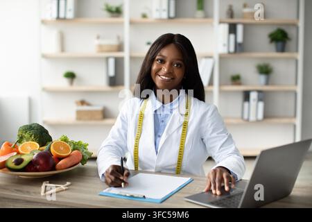 Happy Black Female Nutritionist Writing Meal Plan For Clients In Office, Healthcare Concept Stock Photo