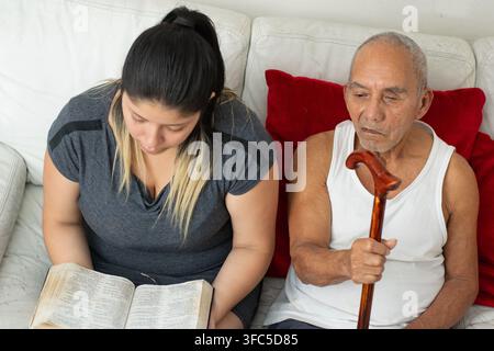 Granddaughter reading book next to elderly grandfather holding cane in supportive home setting Stock Photo