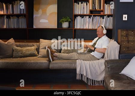 Senior Asian man reclining on right leather sofa, using laptop and wearing headphones, copy space Stock Photo