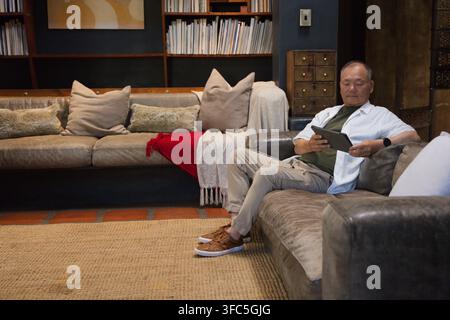 Senior Asian man sitting on right sofa in lounge holding tablet with bookshelves behind, copy space Stock Photo