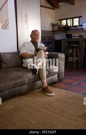 Senior Asian man sitting on brown leather sofa reading paperback book near kitchen counter Stock Photo