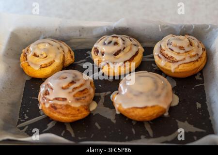Close-up of freshly baked cinnamon rolls covered with sweet icing on a parchment-lined baking tray. Homemade pastry dessert with golden brown swirls a Stock Photo
