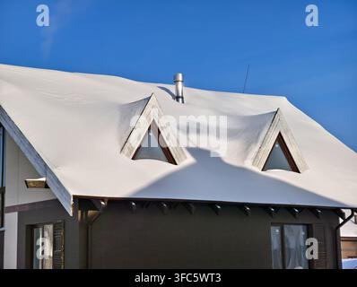 The roof of a cottage covered with snow in winter. A shadow from a neighboring building with a chimney emitting smoke is visible on the roof. Stock Photo