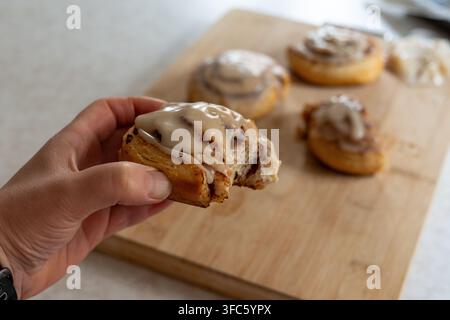 Close-up of a hand holding a partially eaten homemade cinnamon roll covered with sweet icing. Soft layered pastry with visible cinnamon filling, with Stock Photo