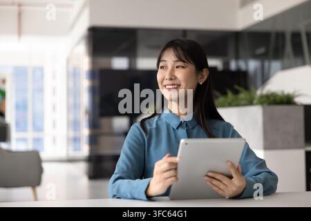 Smiling Asian woman holding tablet at workplace, looks away thoughtfully Stock Photo