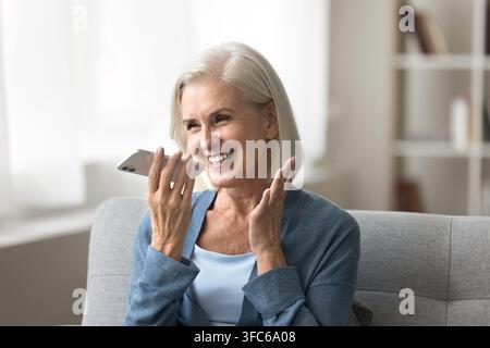 Cheerful elderly woman hold smartphone by mouth record vocal message Stock Photo