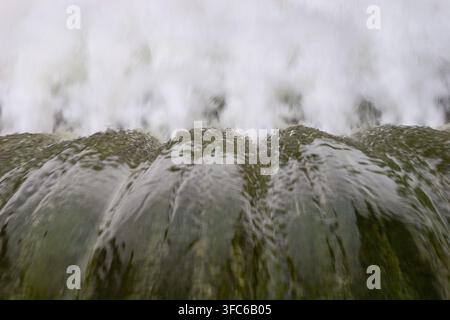 Fast-moving water pouring over a concrete dam edge, creating smooth crests and white foam below. Stock Photo