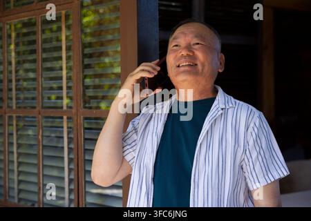 Senior Asian man holding phone to right ear, smiling, looking up on porch by louvers, striped-shirt Stock Photo