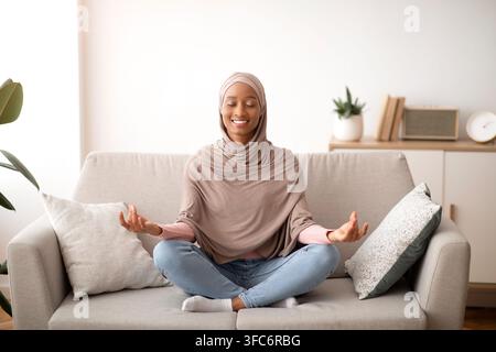 Stress management concept. Peaceful young lady in hijab meditating with closed eyes on sofa at home Stock Photo